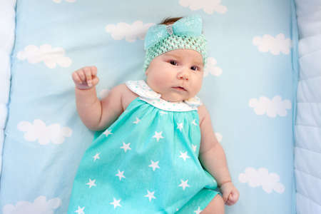 Sweet little baby lying bed with opened eyes. She in blue dress with white stars, bow on head, top...