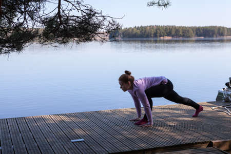 Woman doing yoga exercise on wooden pier on the lake shore at morning time