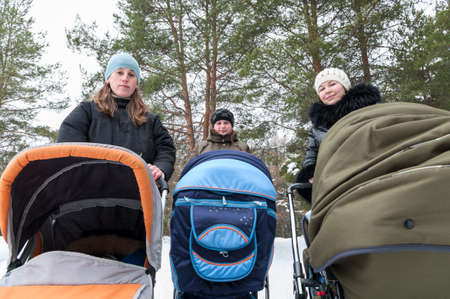 Three young Caucasian mothers walking with strollers in winter forest, portrait, looking at camera...