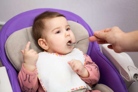 Baby boy first feeding, little child sitting in chair in domestic room, female hand with spoon