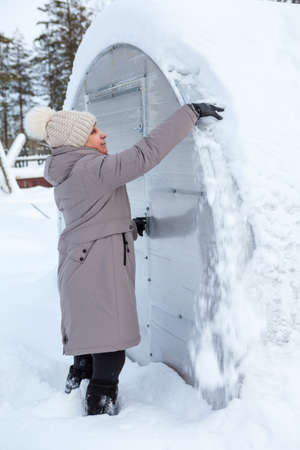 Middle age woman trying to clear the snow away from roof of plastic greenhouse at winter seasonの写真素材