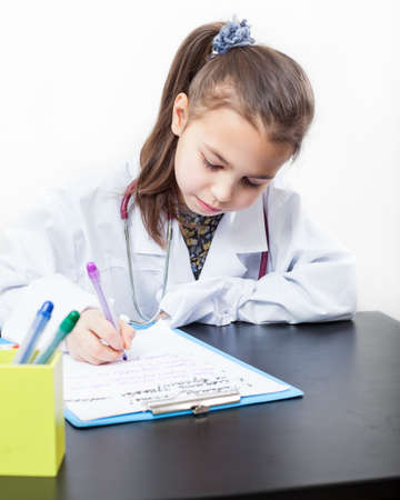 Cute preschool girl playing doctor, dressed uniform sitting at the table and writing in clipboard, white backgroundの写真素材