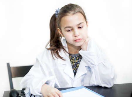 Sad school girl in a white medical coat sits at a table, white backgroundの写真素材