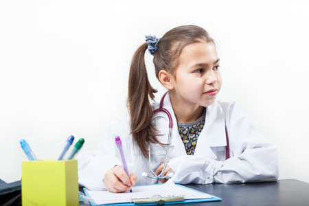 Preschool age girl playing doctror character, white lab coat, sitting at the desk, white background