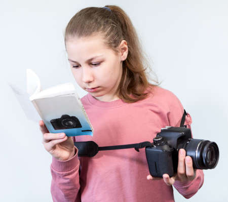 Teenage Caucasian girl reading user manual of DSLR camera, learning photography, a grey background