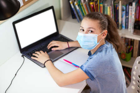 Schoolgirl studying at home wearing medical mask, concept of children distance eduction during...