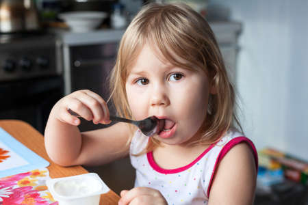 Portrait of young funny girl with spoon in mouth, sitting at the table in domestic kitchenの写真素材