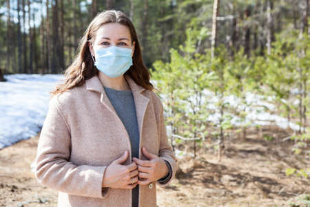 Attractive young Caucasian woman wearing beige coat and medical mask standing in spring forest, portrait with copyspaceの写真素材