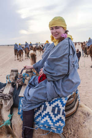European woman dressed long sand protective dress and headscarf sitting on the camel while riding in Sahara desert in Tunisia, Africaの写真素材