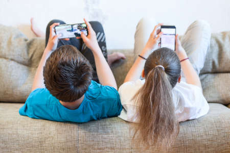 Sister and brother using their smartphones while lying back on a couch at homeの写真素材