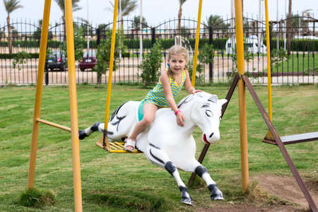 Little Caucasian girl sitting on harseback of children swing, swinging on playgroundの写真素材