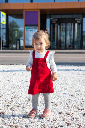 Caucasian toddler girl weraing red dress walking on white pebbles on the street