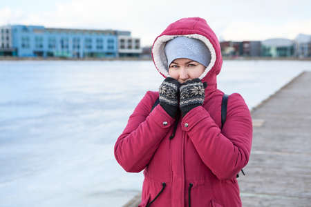 Adult woman wearing warm coat with hood on head and knit mittens standing on quay of a frozen lake
