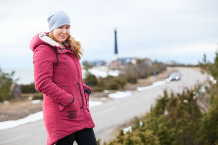 Woman dressing warm red coat standing on hill against lighthouse on sea cape, travel at winter seasonの写真素材