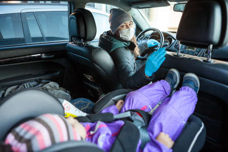 Woman a driver in safety mask and blue gloves sitting on driver chair, looking back at her baby in a chairの写真素材