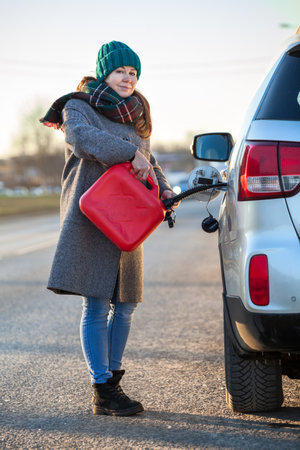 Smiling woman driver pouring diesel in tank of her suv from red plastic can, an accident with empty car-tank on the road at winter seasonの写真素材