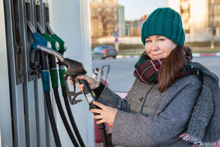 Portrait of woman in winter clothing with gas nozzle in hands, petrol station, looking at camera, cold seasonの写真素材