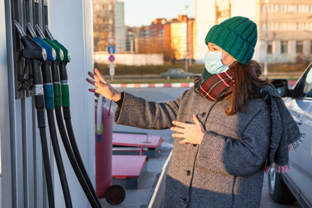 A woman with protective mask chooses brand of fuel at a gas station for a car. Warm clothes, winter seasonの写真素材