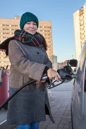 Happy middle age adult woman holding fuel pump by car at self-service gas station in city