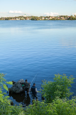 Two adult fishermen catching fish in freshwater lake, town is in the distance
