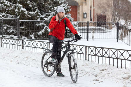 Bicyclist call with smartphone while standing with bike on snowy urban street, riding on bicycle at...