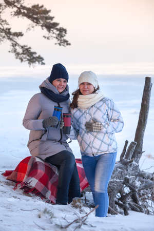 Two girlfriends have winter picnic on snowy sea coast, Caucasian females sitting on plaid covered...