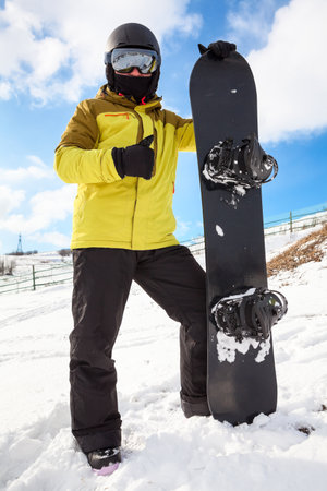 Snowboarder man with snowboard standing on mountain slope, holding his board, wearing mirrored glasses, thumb upの写真素材