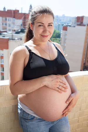 Beautiful pregnant woman on the balcony wearing jeans and underwear holding her tummy, Caucasian femaleの写真素材