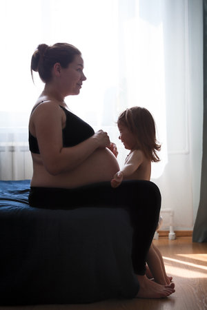 Little daughter happy with pregnant mother, embracing her tummy and laughing, domestic dark room with bright windowの写真素材