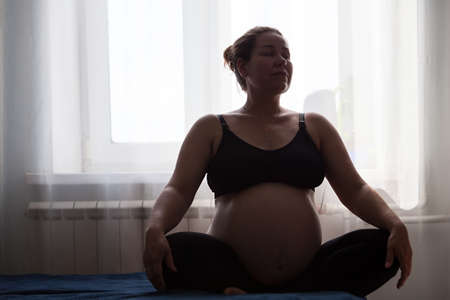 Pregnant woman sitting on her bed in bedroom, lotus pose, resting or meditating in dark room with bright windowの写真素材
