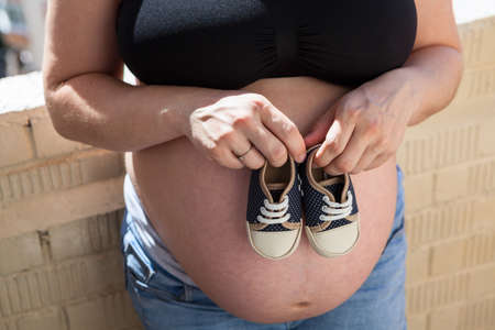 Pregnant woman holds small shoes near her belly, close up viewの写真素材