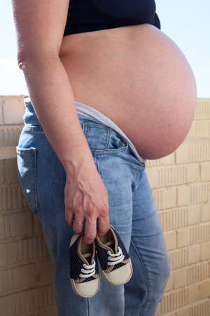Small baby shoes in Caucasian female hand, side view with big pregnant belly, unrecognizable personの写真素材