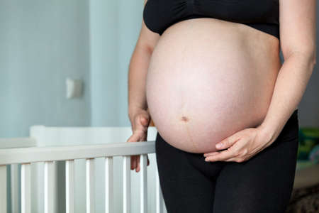 Close up view at pregnant woman big belly next to white baby cot in a bedroom, unrecognizable Caucasian femaleの写真素材