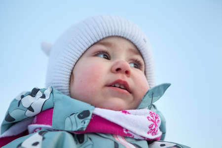 Close up low angle portrait of little child wearing warm hat and scarf, read frozen nose with cheeks, winter season. Caucasian girlの写真素材