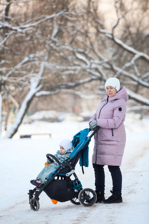 Caucasian mother and her baby in a carriage walking in city park at winter, cold weatherの写真素材