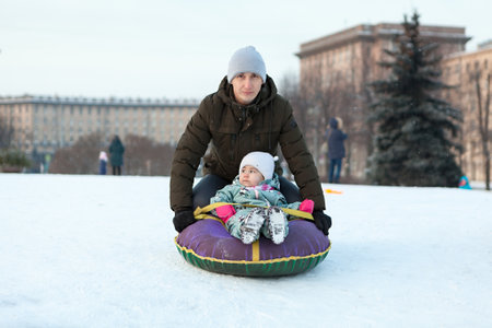 Father push her child from snowy slope, kid sitting in an inflatable toboggan, winter season is in Russian cityの写真素材
