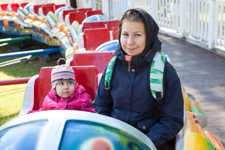 Teenage girl with her little sister riding train on playground in amusement park, sitting together in cabin, autumnの写真素材