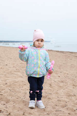 Small girl blows soap bubbles while standing on empty sandy beach of coastline, autumn seasonの写真素材