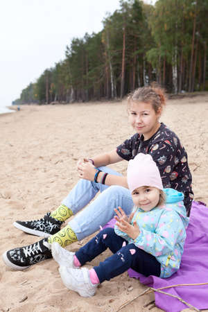 Two and twelve years old children sitting together on empty sandy beach, sisters with different agesの写真素材