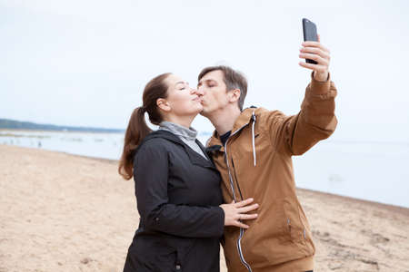 Young happy couple in love kissing and making selfie at the beach, cold season, Caucasian peopleの写真素材