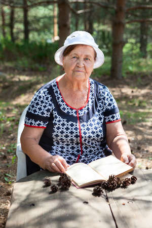 Old lady reading book sitting at the wooden table in a summer forest, looking at cameraの写真素材