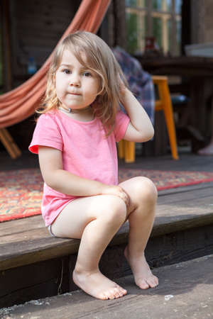 Caucasian little girl wearing pink t-shirt sitting on wooden stairs on patio, summer seasonの写真素材