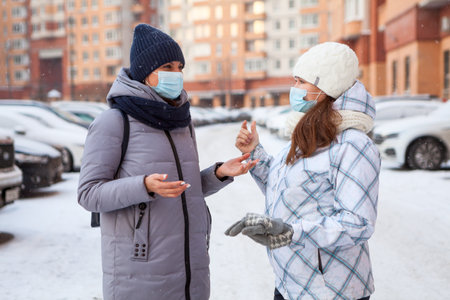 Happy Caucasian women talking and laughing in snowy urban street, females wearing medical masks with winter clothingの写真素材