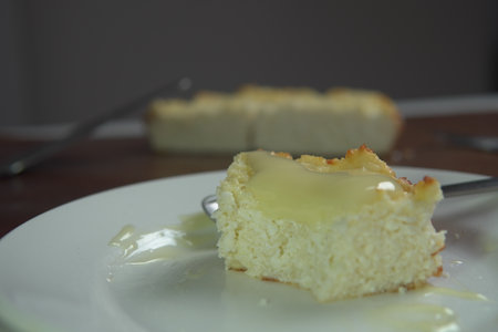 appetizing slice of homemade cottage cheese casserole topped with flowing condensed milk on white plate with fork bite and whole casserole in soft backgroundの写真素材