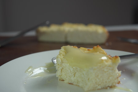 appetizing slice of homemade cottage cheese casserole topped with flowing condensed milk on white plate with fork bite and whole casserole in soft backgroundの写真素材