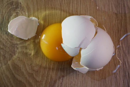 cracked raw egg with bright yellow yolk and spilled white lying on wooden surface surrounded by broken eggshell fragments closeup viewの写真素材