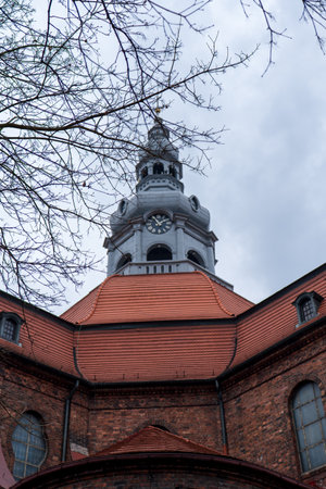 Upward view shows brick walls, copper red roof, and a baroque clock tower in Katowice, Poland, framed by bare spring branches under cloudy sky in soft daylightの写真素材