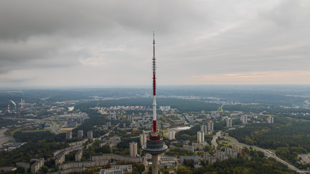 Drone view of the Vilnius Television Tower in Lithuania, featuring its red and white design, urban buildings, forested areas, and an overcast sky.の写真素材