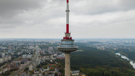 The Vilnius TV Tower stands tall with its red and white antenna, surrounded by urban buildings, dense forests, a winding river, and an overcast sky.の写真素材