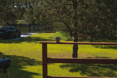 A grassy yard with trees, a wooden railing holding a coffee mug, a car on a gravel driveway, and a charcoal grill in a private countryside setting.の写真素材